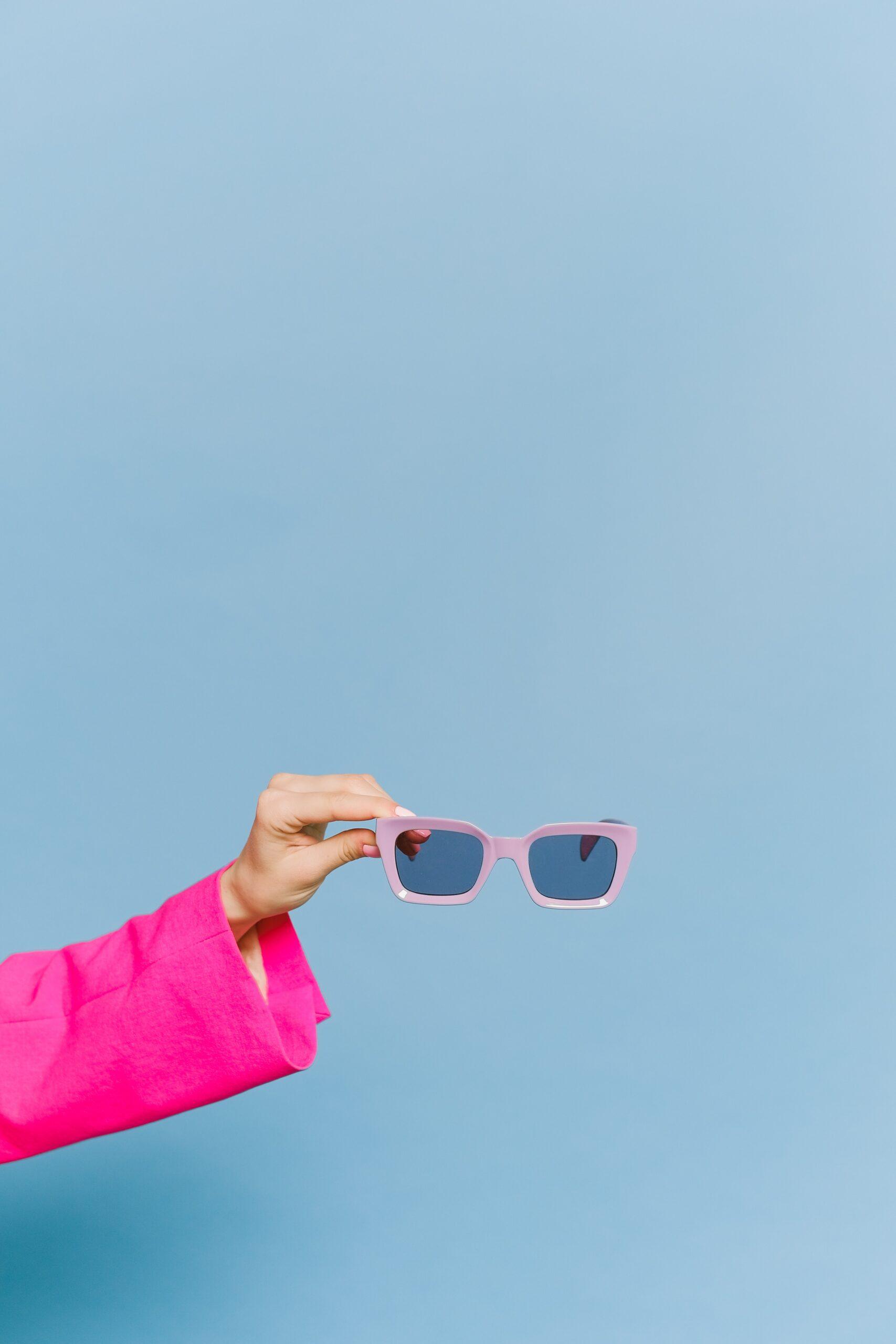 Hand in Pinker Klamotte hält rosa Sonnenbrille vor blauem Hintergrund