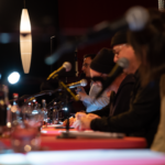 Blick auf die Tische der Lesenden in der Live-Lesung im Finale vom Drehbuchpreis Schleswig-Holstein: Viele Mikrofone, Lesende, Drehbücher und Wassergläser.