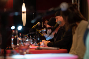 Blick auf die Tische der Lesenden in der Live-Lesung im Finale vom Drehbuchpreis Schleswig-Holstein: Viele Mikrofone, Lesende, Drehbücher und Wassergläser.