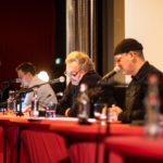 Lesende im Finale vom Drehbuchpreis Schleswig-Holstein: Fokus auf Oliver E. Schönfeld, der mittig im Bild sitzt, und konzentriert mitliest.