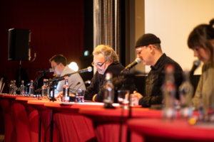 Lesende im Finale vom Drehbuchpreis Schleswig-Holstein: Fokus auf Oliver E. Schönfeld, der mittig im Bild sitzt, und konzentriert mitliest.