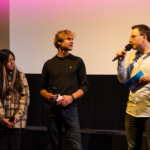 Jasmin Luu und Friedrich Tiedtke stehen vor der Leinwand im Finale vom Drehbuchpreis Schleswig-Holstein und sprechen mit Moderator Horst Hoof.