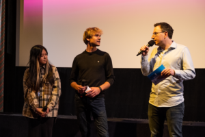 Jasmin Luu und Friedrich Tiedtke stehen vor der Leinwand im Finale vom Drehbuchpreis Schleswig-Holstein und sprechen mit Moderator Horst Hoof.